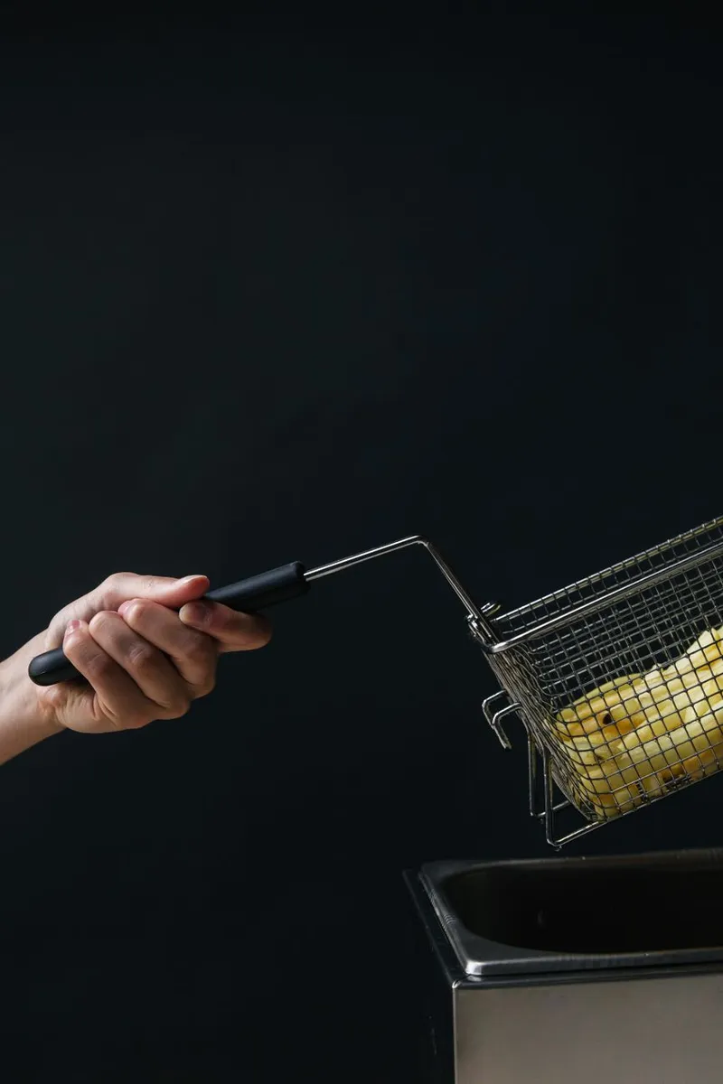 Close-up of a hand holding a fry basket with potato slices over fryer against a dark background.